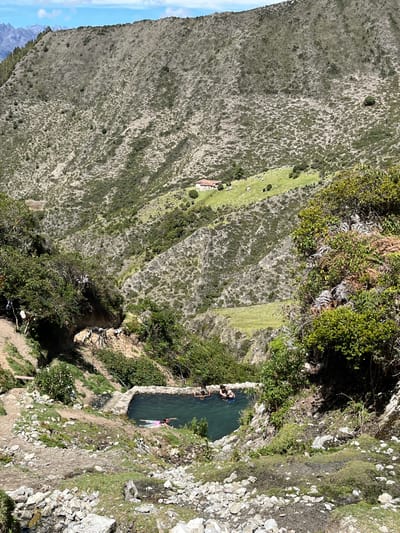 The image shows a natural hot spring pool in a mountainous, rural area. The pool, constructed with stone borders, is filled with dark blue water and has a few people swimming in it. One person in the foreground is swimming on their back wearing a pink swimsuit. The surrounding environment is rugged and natural, with steep, vegetated slopes and rocky terrain. In the distance, a small house with a red roof is visible on a grassy hillside. The sky above is blue with scattered white clouds. The location is identified as a hot spring in Mucurubá, Venezuela.