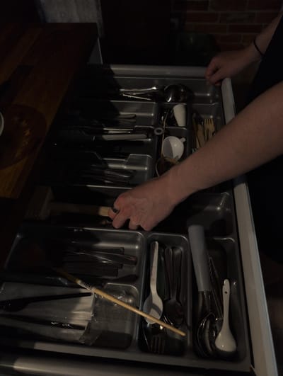 A person's arm and hand are reaching into an open cutlery drawer. The drawer is divided into compartments, holding various kitchen utensils. In the foreground, a wooden mallet rests in one of the compartments, with the person's hand reaching towards it. To the right, a bamboo skewer or chopstick is partially visible, propped up by other utensils. The drawer is filled with a variety of spoons, forks, and other miscellaneous items, some of which are still in plastic packaging. The background shows a glimpse of a brick wall, suggesting an indoor setting. The lighting is dim, casting shadows and highlighting the metallic sheen of some of the utensils. The overall scene depicts someone interacting with the contents of a kitchen drawer. There is no visible text.