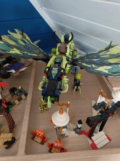 A Lego dragon model is the central subject, positioned on a wooden surface. The dragon is predominantly lime green with black and blue accents on its body, and features large, camouflage-patterned wings in shades of green, yellow, and black. It has black horns and what appear to be glowing red eyes. Surrounding the dragon are various smaller Lego builds.

To the left of the dragon, there is a small Lego boat or raft with purple and blue elements. In front of the boat, a small Lego scene includes a ladder, a black scorpion figure, and other brick-built structures.

In the foreground, to the right of the dragon, are three small, orange Lego figures with angry expressions, resembling crabs or stylized creatures. Further to the right, a more complex Lego structure is visible, featuring white and gray elements with red accents, including a black robotic-looking piece and what appears to be a small white tower with a golden flag. A black and red Lego structure with white steps is also present.

The setting appears to be an indoor display or play area, with the wooden surface suggesting a shelf or tray. The background is out of focus, showing hints of blue and white. No specific time of day or weather conditions can be determined. The overall scene depicts a toy display, possibly part of a larger Lego collection or play scenario. There is no visible text in the image.