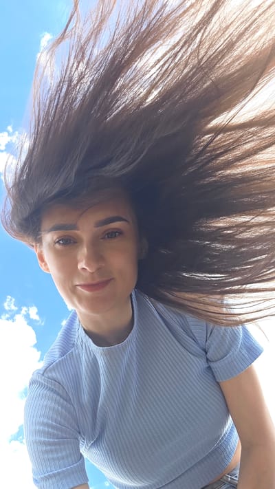 A young woman with long, dark brown hair is captured from a low angle against a bright blue sky with scattered white clouds. Her hair is being blown upwards and to the right by the wind, framing her face. She is wearing a light blue, ribbed short-sleeved t-shirt. Her gaze is directed upwards and slightly to the left, and she has a faint smile. The scene appears to be outdoors, possibly in a park or open area, given the sky and clouds. The location is identified as Дарьино, Russia.