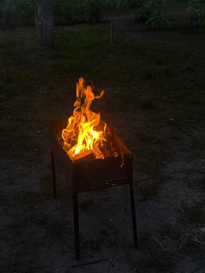 A close-up, eye-level, outdoor shot captures a metal barbecue grill with active flames in Velyka Cherniavka, Ukraine. The grill, positioned on a dark, uneven ground surface with sparse vegetation and some larger stones, is filled with burning wood, emitting bright orange and yellow flames that flicker upwards. The background is dark and out of focus, with hints of greenery, possibly from plants or trees, and a vertical, textured element that appears to be a tree trunk. The overall lighting suggests it is dusk or nighttime.