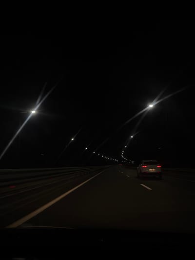 The image depicts a nighttime view from the perspective of a vehicle traveling on a highway. The highway is illuminated by streetlights positioned on tall poles, casting bright beams into the darkness. A white SUV is visible ahead, traveling in the same direction. The road is marked with white lines, and a concrete barrier runs alongside the highway on the left. The scene suggests a drive through a rural or suburban area, given the relative darkness and the presence of only a few other vehicles. There are no discernible landmarks or specific architectural features that would allow for precise identification of the location within Lavrovka, Russia. The image is consistent with a typical highway scene at night.