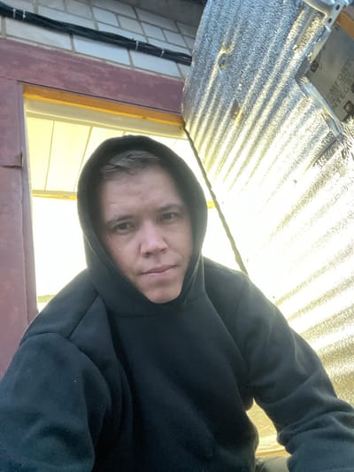 A head-and-shoulders selfie shows a person with short, light brown hair wearing a black hooded sweatshirt. The person is looking directly at the camera with a neutral expression. The background is out of focus, but appears to be outdoors with sunlight reflecting off a textured surface. To the left of the person is a dark red, wooden beam and a brick wall. To the right is a bright, corrugated material with strong diagonal lines of light and shadow. The person is positioned in the foreground, with the background suggesting an exterior of a building, possibly under construction or renovation. The location is identified as Veteran-2, Russia.