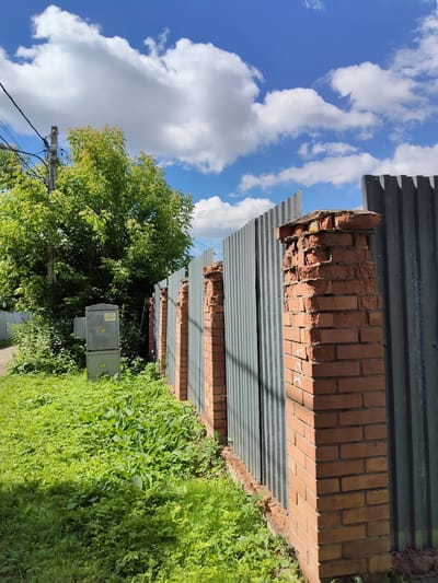 The image depicts an outdoor scene on a sunny day, with a bright blue sky dotted with fluffy white clouds. A corrugated metal fence runs diagonally across the frame, supported by brick pillars. Some of these pillars appear weathered and damaged, with bricks missing or dislodged at the top. Lush green grass and various plants grow abundantly along the base of the fence and in the foreground, creating a slightly overgrown appearance.
To the left of the fence, a grey electrical box is visible, mounted on a pole and surrounded by greenery. The box has two yellow triangular warning signs with exclamation marks. Further to the left, a dirt path leads away from the viewer, disappearing behind dense green foliage and trees. Electrical wires are strung overhead, supported by a utility pole.
The overall setting appears to be a rural or semi-rural area in Венюково, Russia. The strong sunlight suggests it is daytime, likely late morning or afternoon. The scene conveys a sense of an undeveloped or perhaps neglected outdoor space, with elements of both nature and man-made infrastructure. No people are visible in the image. The visible text on the electrical box is a warning label, likely indicating an electrical hazard.