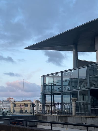 The image shows a partial view of a modern glass and steel building in La Coruña, Spain, taken from a low angle. A large, triangular grey overhang is visible on the left, supported by a cylindrical column. Adjacent to it is a multi-story glass structure with a visible interior, reflecting the exterior environment. Iron railings separate the building from the foreground. The background shows a cityscape with older buildings, their facades visible through the reflections and glass of the modern structure. The sky is overcast with grey and pink-tinged clouds. The image has been rotated 90 degrees counterclockwise.