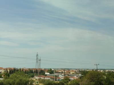 The photograph captures a wide-angle view of a suburban or semi-rural area. In the foreground, lush green foliage dominates, obscuring the lower portion of the scene. Beyond the trees, a residential area unfolds, characterized by buildings with terracotta-tiled roofs, interspersed with cypress trees and other vegetation. Several power lines crisscross the mid-ground, supported by tall, lattice-style electricity pylons. In the distance, a hazy horizon suggests the presence of more buildings and possibly a coastline. The sky is a pale blue, with wispy clouds streaked across it, indicating a daytime setting. The overall impression is one of a quiet, everyday landscape.
