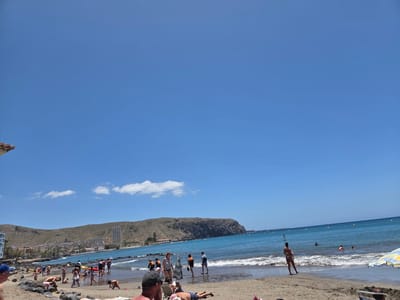 This image shows a crowded beach on a sunny day in Chayofita, Spain. The beach is sandy, with people sunbathing, swimming, and playing. Buildings are visible in the distance, nestled along the coast at the base of a large hill. The sea is blue, with some small waves approaching the shore. Several people are wading in the water along the coastline.