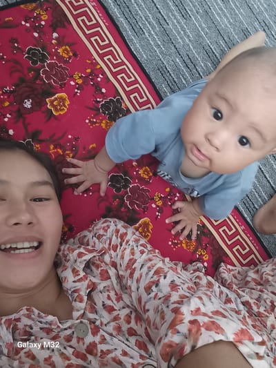 A high-angle, close-up shot shows a woman and a baby lying on a red patterned mat. The woman is in the foreground, smiling at the camera, her face taking up the left side of the frame. She has dark hair and is wearing a floral print top. To her right, a baby wearing a blue shirt is looking up and to the side. The baby's hand is resting on the mat near the woman's head. The mat has a red base with a floral pattern of dark red, yellow, and black flowers. A decorative border with a Greek key pattern runs along the top edge of the mat. The background consists of a grey, textured surface, possibly a rug or carpet. The scene appears to be indoors, in a domestic setting.
