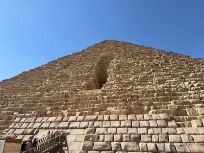 This media file features a low-angle, upward-facing view of a large, ancient stone pyramid under a clear, bright blue sky. The pyramid is constructed from numerous large, rectangular stone blocks, weathered and varying in color from light tan to brown. The surface texture of the pyramid is rough and irregular, with many visible seams between the blocks. A prominent, dark, rectangular opening, likely an entrance or passage, is visible near the top of the pyramid, slightly off-center to the left.

In the lower left quadrant of the image, a group of people are visible on and around a wooden staircase leading up the side of the pyramid. There are approximately seven to eight individuals, some of whom appear to be tourists. They are dressed in casual, light-colored clothing. Their presence suggests an archaeological site or tourist attraction.

The lighting indicates it is daytime, likely midday or early afternoon, given the strong sunlight casting shadows and illuminating the stone surfaces. The clear blue sky suggests fair weather conditions. The presence of the pyramid itself strongly implies a location of historical significance, consistent with the provided location context of Kafrat Nassar, Egypt, known for its ancient Egyptian monuments. The overall mood is one of awe and historical exploration. There is a small, wooden sign with text in the bottom left corner of the image, but the text is too small and blurry to be legible.