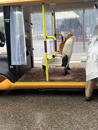 The image shows the exterior of a yellow and brown autonomous electric vehicle in Nagakute, Japan. The vehicle has a clear plastic curtain on the side and a tan seat with a brown teddy bear on it. A person in a white rain suit and tan pants is standing to the right of the vehicle. The ground is wet, indicating it is raining. In the background, there are trees and a white tent. The vehicle has the text "APM NB" on the side. The scene appears to be a parking area or street in a somewhat urban or park-like setting.