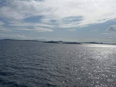 This image depicts a wide, dark blue sea under a cloudy sky. In the distance, a chain of small, dark, low-lying islands or landmasses stretches across the horizon, appearing silhouetted against the lighter sky. The water is choppy with a moderate swell, and sunlight glints off the wave crests in the mid-ground and right side of the image. The sky is filled with a mix of white and gray clouds, with patches of blue visible. The overall impression is of a vast, open seascape. The scene is likely a view of the coast near Nordsolvær, Norway, a known archipelago characterized by similar island formations and proximity to the ocean. There are no people or identifiable man-made objects visible in the image, and the focus is entirely on the natural environment.