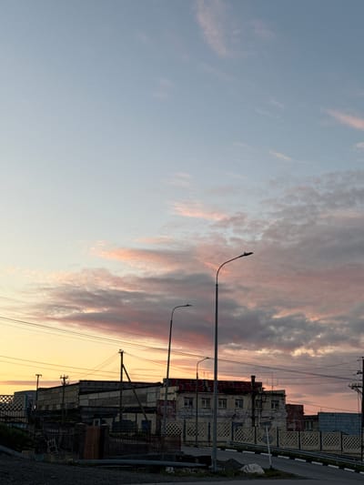 The image depicts a twilight scene in Anadyr, Russia, focusing on an industrial or utilitarian area. The sky is a gradient of pale blue at the top transitioning to hues of orange and pink near the horizon, indicative of sunset or sunrise. Scattered clouds with pink and grey tones are visible against the lighter sky.

In the foreground, a paved road curves to the right, with a gravel shoulder on the left. A white line marks the edge of the road. There are traffic barriers and a simple fence. A white plastic bag rests on the roadside.

Dominating the midground are several low-rise buildings, appearing somewhat dilapidated. They are constructed with concrete blocks and have visible windows. Some of the buildings display graffiti in red paint. Electrical poles and wires are strung across the scene, and there are streetlights. A fence with a diamond pattern runs in front of some of the buildings.

In the background, more buildings and structures are visible, blending into the twilight. The overall impression is one of a functional, perhaps slightly neglected, urban or industrial landscape under a colorful sky. There are no people visible in the image.