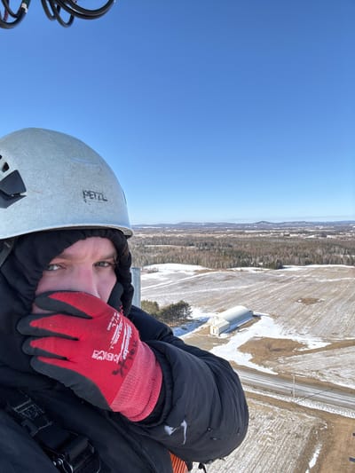 A person is seen from an elevated perspective, wearing a light grey Petzl helmet, a black hooded jacket, and red Milwaukee work gloves (identified by "Milwaukee Cut Level 1" text). The gloved right hand covers the lower half of the face, leaving only the eyes and forehead visible. Black cables are partially visible in the upper left corner. The background reveals a panoramic view of a snow-dusted rural landscape under a clear, bright blue sky. Below the vantage point, fields show patches of snow and exposed brown earth, indicating winter or early spring conditions. A paved road cuts through the mid-ground, flanked by a long, arched-roof building and a smaller rectangular structure. Beyond these, dense forests, comprising both deciduous and coniferous trees, extend towards distant low-lying hills or mountains on the horizon. The scene is located in Easton, Maine, United States of America.