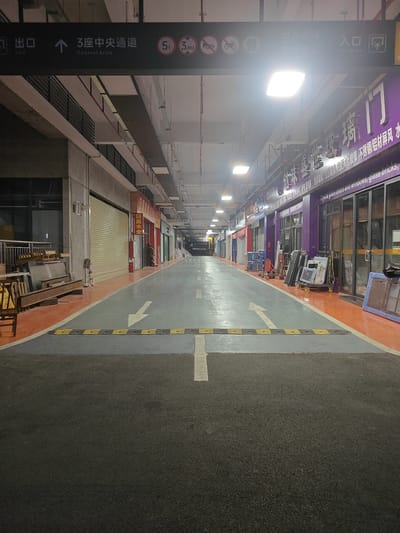 The image shows a dimly lit, long, empty indoor market or storage facility in Nanhai District, China. The perspective is from a ground-level view looking down a wide, paved pathway. On the left side of the pathway, there are closed storefronts with metal roll-up doors and some exposed metal beams and wooden chairs. On the right side, there are also storefronts, some with glass doors and windows displaying various items, including what appears to be glass doors and windows, and stacks of materials. Overhead, a sign in Chinese and English indicates "Central Aisle" and shows directional arrows, weight limits (5t), and height limits (3.8M). Further down the aisle, more storefronts are visible, and the lighting fixtures are prominent on the ceiling. The pathway is marked with white arrows and a speed bump. The overall environment appears to be a commercial or industrial space that is currently closed or unoccupied.