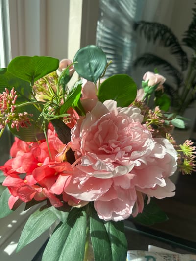 This media file displays a close-up view of a floral arrangement, likely artificial, situated indoors near a window. The arrangement features large, delicate pink peonies, smaller coral-colored blooms, and various green foliage. The lighting suggests it is daytime, with sunlight casting distinct shadows across the scene, creating a bright and airy atmosphere. The background is softly blurred, but hints of Venetian blinds or a similar window covering are visible, indicating an interior setting. The overall mood is serene and decorative. There is no discernible text within the image.