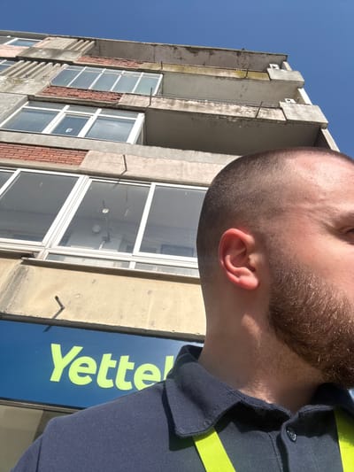 An upward-angled outdoor photograph captures the upper facade of an aging multi-story concrete building, a partial view of a male subject in the foreground, and a clear blue sky. The male subject, positioned in the lower right foreground, displays the right side of his head, including a short haircut, a beard, and his ear. He wears a dark blue collared polo shirt, with a neon yellow-green strap visible over his right shoulder. The building, consistent with structures found in the city of Levski, Bulgaria, features a weathered concrete exterior with signs of discoloration and minor structural wear. Multiple windows with white frames and internal blinds are visible, interspersed with sections of exposed red brickwork. Concrete balconies are present on the upper levels. Through a central window, an interior light fixture and a multi-element antenna structure are discernible. Below the windows, a blue commercial sign prominently displays the bright yellow-green text "Yettel." The sky above the building is uniformly clear and blue, indicating daytime conditions.