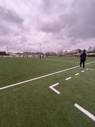 The image displays an artificial turf football pitch in Montigny-lès-Metz, France, under an overcast sky. In the immediate foreground, a person stands with their back to the camera, dressed in a dark hooded jacket, dark trousers with a white vertical stripe, and white athletic shoes. They are positioned to the right of a solid white sideline marking, with additional white dashed lines and a white arrow-shaped marking visible on the green turf.
In the mid-ground, a group of approximately 10-15 individuals, presumed to be football players, are distributed across the field. They are wearing sports kits, predominantly green and red, and appear to be stationary or moving casually, suggesting a practice session or a pause in play. A white football goal is partially visible on the right side of the pitch in the distance.
The background features a long, single-story building with a light-colored facade and a red roof, situated beyond the far side of the pitch. Multiple tall floodlight poles are erected around the field. Deciduous trees, mostly bare of leaves, line the perimeter, indicating late autumn or winter. Residential buildings are visible further in the background beyond the tree line. The overall environment is illuminated by diffuse light consistent with heavy cloud cover.