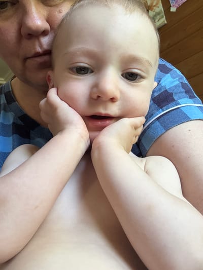 This is a close-up, vertically oriented photo of a baby being held by an adult. The baby, with light skin and light hair, looks directly at the camera with a neutral expression. The baby's right hand is near its mouth. The adult's face is visible in the lower left corner, with their nose and mouth showing. They appear to be resting their head or shoulder on the baby's head.
The baby is wearing a blue and white plaid shirt, and the adult is wearing a similar blue plaid garment, likely pajamas or loungewear. The background consists of a wooden surface, possibly a wall or furniture. The lighting suggests an indoor setting during the daytime. The overall mood is intimate and relaxed. No visible text is present.