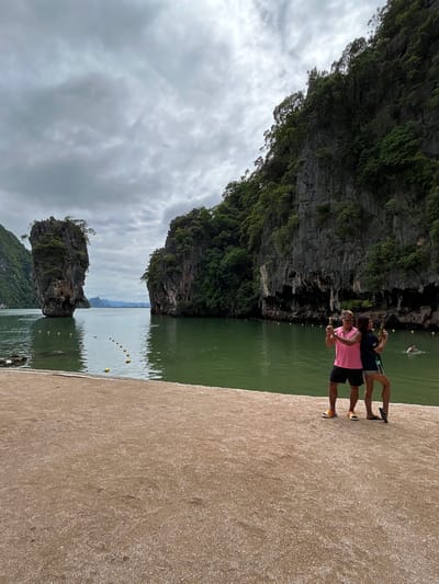 The image captures a scenic view of Phang Nga Bay in Krasom, Thailand, with its iconic limestone karsts. In the foreground, a sandy beach stretches across the bottom of the frame. Two individuals, a man in a pink t-shirt and black shorts, and a woman in a dark t-shirt and light shorts, stand close together on the beach, posing for a photo with water guns. They are positioned in the middle ground, with the water of the bay behind them. The bay itself is a vibrant green, dotted with yellow buoys. To the left and right of the bay, large, jungle-covered limestone karsts rise dramatically from the water. One karst on the left is particularly distinctive, standing tall and isolated. In the distance, the hazy outline of more karsts and mountains can be seen against a cloudy sky. The overall atmosphere is one of a tropical tourist destination.