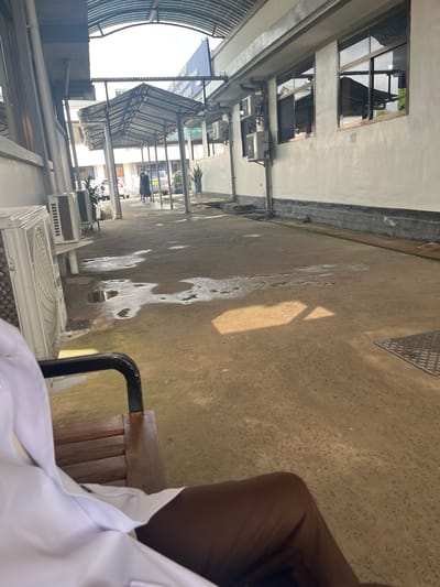 The image shows an outdoor view of a building's exterior in Picha Ya Ndege, Tanzania. The perspective is from someone sitting on a chair near the building. A section of the building on the left has multiple windows and air conditioning units. The ground surface is concrete and appears to have some damp spots. People are walking in the distance under a covered walkway. A leg and part of a white shirt belonging to the person taking the photograph are visible in the foreground. The overall environment suggests an urban setting with a mix of architectural features.