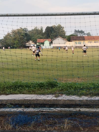The image depicts a soccer game in progress at the "SD Negeri 01 Botok Kerjo" in Botok, Indonesia. Several players are visible on the grassy field, wearing numbered jerseys. A soccer net obscures the view, with overgrown vegetation and concrete structures in the foreground. Houses and trees are visible in the background. The sky is overcast.