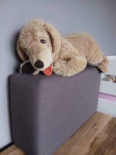 A large, plush, tan stuffed dog with floppy ears and a red tongue sticking out lies on top of a dark grey rectangular ottoman. The ottoman is positioned in the corner of a room, against a light grey wall. To the right of the ottoman, part of a white piece of furniture, possibly a bed or shelving unit, is visible, with some pink material peeking out from inside. The floor in front of the ottoman is made of wood. There are no people or any other discernable animals present in the image. The scene appears to be indoors, likely a bedroom or playroom. The image was taken at an angle looking down at the stuffed dog and ottoman.
