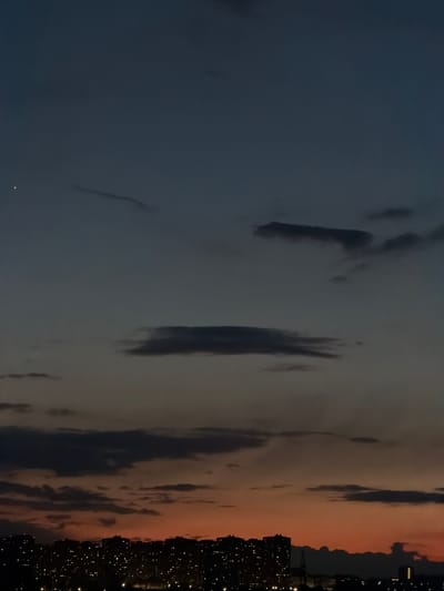 The image shows a night scene of a city skyline, likely taken at dusk or dawn. The sky transitions from a dark blue on the left to a pale orange on the right, with scattered clouds. On the right side of the image, a city skyline is visible, with many illuminated buildings. The bright dots of light are most likely lights from buildings. Given the limited information and the general nature of the image, determining if this is Olginskaya, Russia, or the specific location within the city is impossible.