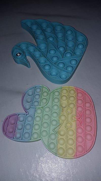 Two pop-it fidget toys are shown on a white, textured surface. The top toy is blue and shaped like a swan or peacock. It has a beak, an eye, and rows of circular bubbles covering its body. The bottom toy is shaped like a mouse or similar cartoon character and is colored with a gradient of pastel shades, including purple, blue, green, yellow, and pink. This toy also features multiple rows of circular bubbles. The background is dark and out of focus. The image is a still photograph. The location of Sedayu, Indonesia, is not discernible from the image itself.