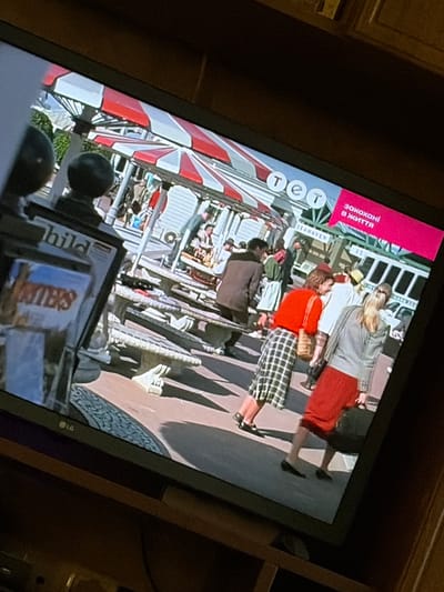 The image shows a television screen displaying a scene of people in what appears to be an outdoor market or fair. There are several people visible, some walking and some sitting. In the foreground, there is a television screen displaying an image of a scene with people under a red and white striped canopy. The TV screen has a logo that appears to be "LG". There is also text on the TV screen that reads "SEAHAVEN" and in a pink box "закохані в життя" which translates to "in love with life". The image is taken at an angle, suggesting it is a photograph of a television screen. The location in the city of Osolynka, Ukraine, is not depicted in this image.