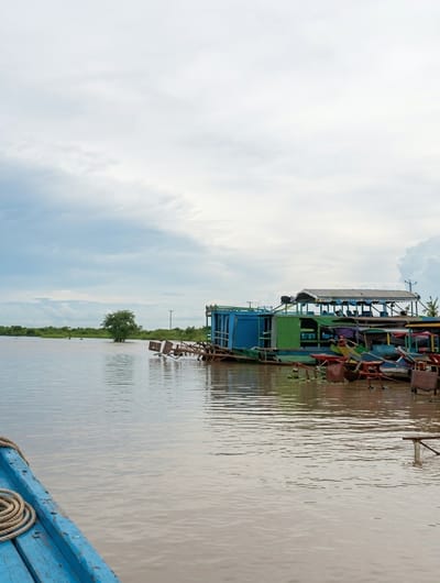 The image depicts a wide, calm body of water, likely a river or lake, under an overcast sky. In the middle ground, a collection of houseboats and possibly barges are moored together. One prominent houseboat is painted in vibrant blue and green sections and features a multi-tiered roof structure covered with a striped tarp. Other boats, some with colorful elements, are visible alongside it. On the left side of the image, a portion of a blue wooden boat, likely the vantage point for the photograph, is visible in the foreground, with a coiled rope on its deck. The background shows a green, tree-lined shore with a single prominent tree in the distance. The overall scene suggests a tranquil, possibly residential or commercial, waterfront area in a Southeast Asian setting, consistent with locations like Soutr Nikom, Cambodia, known for its floating villages and waterways. There are no visible people in the frame.