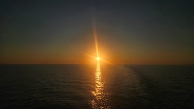 The image displays a serene ocean scene at sunset, with the sun appearing as a bright, radiant orb positioned at the center of the horizon. The sun's rays create a shimmering reflection on the water, stretching outwards towards the viewer. The sky transitions from a deep, dark blue at the top to a warm, gradient of orange and yellow near the horizon, indicating the fading light of day. The ocean itself appears calm with subtle ripples on its surface. The location is specified as Unknown, Sweden. There are no visible people, structures, or specific landmarks within the frame, emphasizing the vastness and natural beauty of the seascape.
