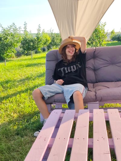 In Nechaiane, Ukraine, a young person wearing a straw hat, a black t-shirt, and denim shorts is sitting on a cushioned sofa outdoors. The sofa is made from stacked wooden pallets painted pink and purple. The person is leaning back on the sofa, with their left arm raised and bent behind their head, and their tongue is sticking out playfully. The scene is set in a grassy area with rows of young trees, likely an orchard, under a clear sky with a large white canopy overhead. The lighting suggests it is a sunny day.