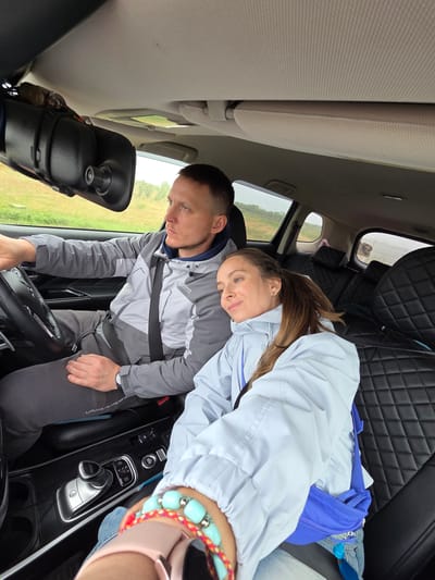 The image captures two people, a man and a woman, inside a car, likely in Бабстово, Russia, during an overcast daytime.
The man is seated in the driver's seat, wearing a two-toned grey jacket with "FAVORITE" and "SPORTSWEAR" visible on it, and a seatbelt across his chest. He looks down intently, possibly at the steering wheel or controls, with a focused expression. A watch is visible on his left wrist.
Leaning against the man's shoulder and looking towards the camera is the woman. She has long brown hair and wears a light blue or light grey jacket. Her expression is soft and gentle. On her right wrist, she wears a multi-colored beaded bracelet, predominantly teal, and a pink smartwatch. A bright blue bag is tucked beside her.
The car's interior features black, diamond-quilted leather seats and a dashboard with an automatic gear shift (P, R, N, D, M settings are visible) and various control buttons. A dashcam is mounted on the rearview mirror. Through the car windows, a green, grassy landscape is visible under what appears to be a cloudy sky, indicating overcast weather.