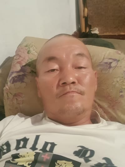 A close-up, selfie-style photograph shows a middle-aged Asian man with a shaved head, lying down. He is wearing a white t-shirt with a dark graphic design. The man is positioned in the foreground, with a pillow patterned with flowers behind him. The background is blurred, but appears to be an indoor setting with textured walls. There is no indication of the specific location within the city of Rassvet, Kazakhstan.