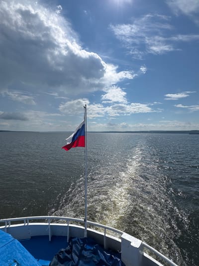 The image captures a view from the stern of a boat sailing on a wide body of water, likely a river or lake, under a bright, partly cloudy sky. The Russian flag is prominently displayed on a flagpole to the right of the frame, its white, blue, and red colors catching the sunlight. The boat's wake stretches out behind it, creating a foamy trail on the dark water, which reflects the sunlight in shimmering patterns. The horizon is lined with a distant, low-lying landmass. The scene appears to be set in the vicinity of Voronovka, Russia, judging by the landscape which is consistent with the region's geography. No people are visible in the image. The overall atmosphere is one of peaceful travel on the water.