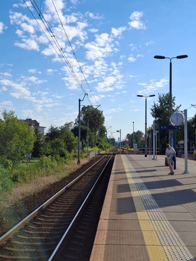 This is an eye-level, medium shot of a train station platform in Ząbki, Poland. The scene is set on a sunny day with a bright blue sky filled with scattered white clouds. The foreground is dominated by a train track to the left, with the station platform occupying the right side of the image.

On the platform, a woman in a light-colored dress and sandals stands facing left, holding a light-colored bag. She appears to be looking down. To her left, a large, round clock is mounted on a post, with its face visible. There are two platform indicators labeled "1 tor" and "2 tor". Further down the platform, another person, dressed in a blue shirt and shorts, is walking away from the camera. The platform surface is paved with textured tiles and features tactile paving for visually impaired individuals.

The background shows lush green trees and some buildings. Overhead, power lines for the electric train are suspended from poles. The train tracks extend into the distance, disappearing under a bridge structure. The overall impression is one of a quiet, functional train station on a pleasant day.