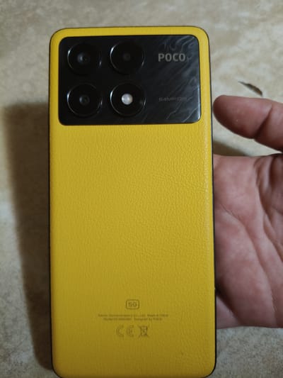 The image shows the back of a yellow POCO smartphone, likely a model with a textured faux-leather finish. The camera module is located in the upper left quadrant of the phone's back and features four circular lenses arranged in a 2x2 grid. The POCO logo is printed to the right of the camera module, and below it, the text "64MP OIS" is visible. Towards the bottom of the phone, a "5G" logo is present, followed by smaller text indicating "Xiaomi Communications Co., Ltd. Made in China" and "Model: 22101316G". The CE mark and a recycling symbol are also visible below the model information. A person's hand, with visible fingerprints, is holding the right edge of the phone, partially obscuring the side. The background appears to be a textured surface, possibly a table or countertop. The scene is indoors, with neutral lighting. The image does not contain any specific visual indicators that would place it in Chornobaivka, Ukraine.