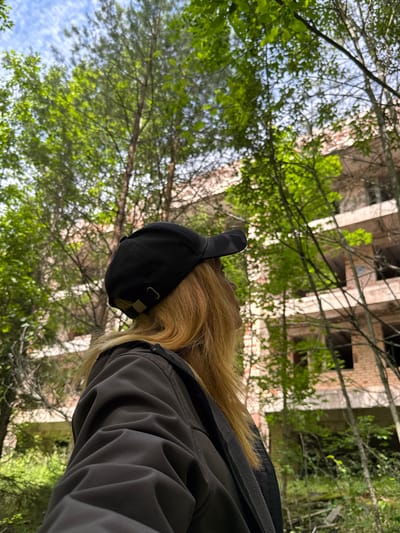 In the city of Interkompleks, Russia, a person wearing a black baseball cap and a dark jacket is viewed from behind, looking towards an abandoned brick building overgrown with trees. The building appears to be several stories high with visible windows and balconies. Lush green foliage surrounds the structure, with sunlight filtering through the leaves, creating a dappled effect on the scene. The sky above is partly cloudy, with patches of blue visible. The overall impression is one of decay and nature reclaiming an urban environment.