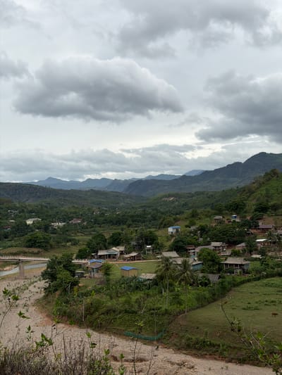 The image depicts a rural landscape in Tà Rụt Commune, Vietnam, under a heavily overcast sky. A wide, rocky riverbed occupies the foreground, with a concrete bridge spanning the water in the middle ground on the left side of the frame. On the right bank of the river and extending into the mid-ground and background is a village composed of several traditional wooden houses with corrugated metal or tiled roofs. The village is nestled among lush green vegetation, including trees and cultivated fields. In the background, rolling hills and mountains covered in dense forest recede into the distance. The overall scene conveys a sense of tranquility and the natural beauty of the Vietnamese countryside. There are no visible people or animals in the image.