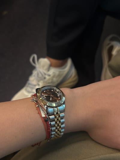 The image shows a person's wrist wearing a two-tone Rolex Datejust watch with a brown dial and fluted bezel. The watch band is two-tone, silver and gold. Adjacent to the watch are two thin, braided bracelets in shades of red and orange, adorned with small charms. In the background, there is a beige sneaker with white laces and a portion of dark fabric, possibly part of clothing. The setting appears to be indoors, likely a casual or personal space. There is no information to suggest that the image was taken in Alessandria, Italy.