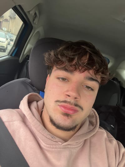 A male subject is depicted in a close-up, front-facing image, seated within the driver's compartment of a vehicle. He exhibits medium-length, dark brown, curly hair, light brown eyes, and fair skin with observable erythema on the forehead and cheeks. Distinct facial hair includes a thin mustache extending to a chin strip. The subject's lips are full. He is attired in a light pink hooded sweatshirt and a dark-colored seatbelt diagonally across his torso. The vehicle's interior shows a dark headrest and a light blue A-pillar. Through the front left window, an exterior scene is visible, consisting of a paved roadway or parking lot with multiple stationary vehicles and a multi-story, light-colored building featuring a balcony. This location is identified as Santa Marta de Penaguião, Portugal, under ambient lighting conditions suggesting overcast weather.