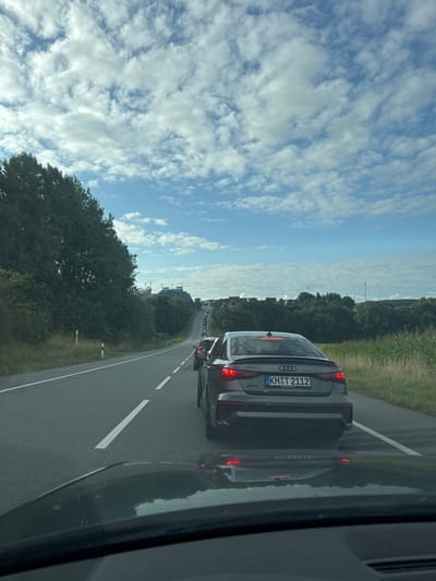 The image depicts a road scene in Hermsdorf, Germany, viewed from the interior of a vehicle. A dark grey Audi RS3 sedan, bearing German license plate KH T 2112, is positioned directly ahead in traffic, with its red LED taillights illuminated. A queue of additional vehicles extends uphill into the distance. The road features a solid white line on the left and a dashed white center line. The left side of the road is bordered by a steep embankment covered with dense green trees and a grassy verge containing a white and black marker post. The right side of the road fronts a field of tall, light green crops, followed by a line of shorter green trees. In the background, on a distant hill to the left, modern buildings with reflective surfaces are visible. To the right in the distance, a series of white wind turbines are present on a ridge. The sky is partially cloudy, with numerous white cumulus clouds scattered across a bright blue expanse. The foreground reveals a dark dashboard or hood and reflections of the Audi's rear on the observer's windshield. No individuals are explicitly visible.