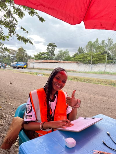An outdoor scene in Usa River, Tanzania, shows a young Black female seated at a light blue plastic table under a red fabric canopy. She is smiling directly at the viewer, giving a thumbs-up gesture with her right hand. Red pigment is present on her forehead and cheekbones. She wears a black and white t-shirt covered by an orange high-visibility vest. Her left hand rests on a clipboard with papers. On the table, to her right, are a white and pink earbud case and several charging cables.

The background reveals a dirt path leading to a paved road. Across the road, a white concrete block wall with a building featuring a rusty corrugated iron roof behind it is visible. Numerous green trees and utility poles with power lines line the horizon. To the far left, a blue flatbed truck covered with a white tarp is on the road, alongside several individuals and a yellow vehicle. The sky is overcast with heavy cloud cover. Green foliage from an overhead tree extends into the upper left corner of the frame.