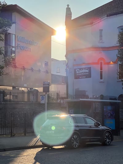 The image captures an outdoor scene in the city of Derry/Londonderry, United Kingdom, likely during golden hour due to the strong sunlight. In the foreground, a dark-colored SUV is parked on the street, facing right. The vehicle's license plate is visible, reading "181-DL-3885". A lens flare from the sun creates a prominent circular light effect over the car. The street has yellow lines painted on it.

Behind the car, a bus stop shelter is situated. A sign on the bus stop pole indicates "Bus Stop" and displays route information. Further back, a black metal fence runs along the edge of a raised area.

Two buildings dominate the background. The building on the left is modern with a light-colored, reflective facade, featuring large windows and prominent silver lettering that reads "Millennium Forum". The building on the right is older, painted a light blue with orange trim. It has arched window frames and a sign for "Rainbow Chinese Restaurant" with operating hours listed as "4pm - 8.30pm".

The sun is positioned high in the sky, between the two buildings, casting long shadows and creating a bright glare that washes out parts of the scene. Trees are visible on the left and right edges of the image. The sky is clear and light blue.