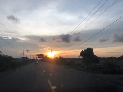 The image captures a road scene in Keudebah, Indonesia, during sunset. A paved road with yellow lane markings extends into the distance, flanked by dark green foliage and vegetation on both sides. Several utility poles with wires are visible along the road. In the foreground, the road surface reflects sunlight, creating blurry circular bokeh effects. Two cars are seen driving on the road, with one white car further ahead and another vehicle further in the distance. The sky is filled with a gradient of colors from pale blue at the top to vibrant orange and yellow near the horizon, indicating the setting sun which is partially obscured by a utility pole and trees on the right side of the frame. The overall atmosphere is serene, characteristic of a rural landscape at dusk.