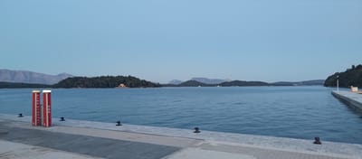 The image captures a coastal scene in the Municipal Unit of Ellomenos, Greece, under soft, diffused light, suggesting dusk or dawn. In the foreground, a paved stone and concrete dock extends from the lower left towards the lower right, featuring several black metal mooring bollards. Two prominent red and white cylindrical pillars stand upright on the dock in the left-center; the left pillar is labeled "FIRE HYDRANT" vertically in white text, and the right pillar is labeled "SOS" vertically in white text. A white pole with a light fixture is visible at the far right end of the dock.

The midground is dominated by a calm body of water with subtle ripples, reflecting the pale sky. Several heavily wooded islands are scattered across the water. On the largest island in the center, a small, light-colored building, possibly a chapel or house, is visible nestled among the trees. Further into the midground and background, additional islands and landmasses covered in dark vegetation extend across the horizon line.

In the distant background, a range of light grey mountains with visible rock formations spans the left half of the image. The sky above is a clear, unblemished pale blue, gradually lightening towards the horizon. No people or active interactions are visible in the scene.