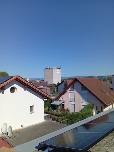 This is a medium-angle, eye-level, daytime, exterior, full shot of a residential area in Murten, Switzerland. The sky is clear and blue. In the foreground, there are solar panels mounted on a rooftop. In the midground, there are several houses with red-tiled roofs. One house is white with a circular window and a small rectangular window. Another house is light pink with white shutters and balconies. Behind these houses, a tall, light-colored concrete building with antennas on top is visible. A Swiss flag is flying from the top of the building, which also has the "Landi" logo on its side. In the background, there are hills and mountains. The scene depicts a typical urban or suburban environment with residential housing and some commercial or industrial infrastructure.