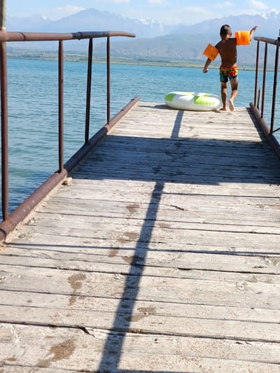 In Kirpichny, Kyrgyzstan, a young boy with orange arm floaties stands on a weathered wooden pier extending into a large body of water. He is wearing multi-colored swim trunks and appears to be in motion, possibly about to jump. A green and white inflatable ring lies on the pier near him. The background features a serene landscape with a vast blue body of water, distant hills, and snow-capped mountains under a clear sky. The pier has a weathered metal railing running along its edge. The sunlight casts long shadows on the wooden planks of the pier.