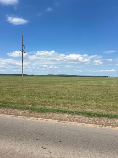 This is a landscape photograph taken on a bright, sunny day in Гудзевіцкі сельскі Савет, Belarus. The scene is dominated by a vast, green field stretching horizontally across the image. The field appears to have been recently mowed, with clear rows visible. In the distance, a dark line of trees forms the horizon, separating the field from the bright blue sky.
The sky is filled with scattered white clouds, some fluffy and others more wispy. A single wooden utility pole, part of a power line, stands prominently in the left side of the frame, silhouetted against the sky. The power lines themselves are visible stretching across the image.
To the right of the field, a rough, unpaved shoulder transitions into an asphalt road that occupies the lower right portion of the image. The road is textured and shows some dark spots, possibly from oil or tar.
The overall impression is one of rural tranquility and an expansive, open landscape. The time of day appears to be late morning or early afternoon, given the bright sunlight and the shadows cast. No people are visible in the image. There is no visible text.
