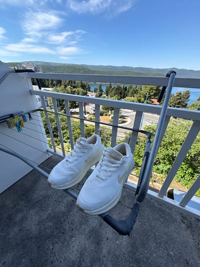 Two white Nike sneakers are drying on a metal clothes drying rack on a balcony. The background shows a view of Neum, Bosnia and Herzegovina, including water, trees, buildings, and a road. Several clothespins are attached to the lower portion of the drying rack.